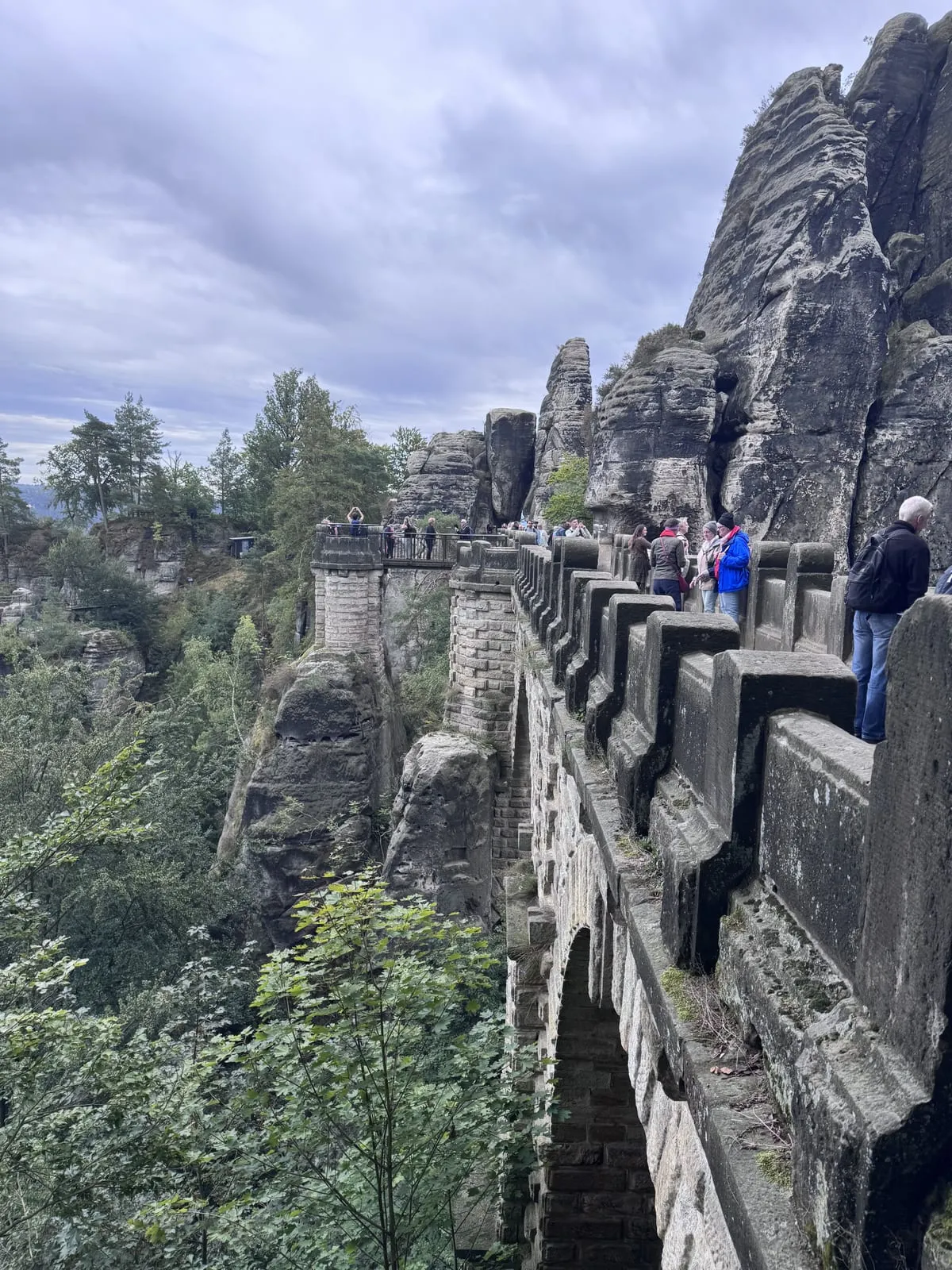 The Bastei Bridge, an arched sandstone bridge with a stone parapet, curving along jagged rock pillars above a forested gorge, with visitors gathered on it under a grey sky.