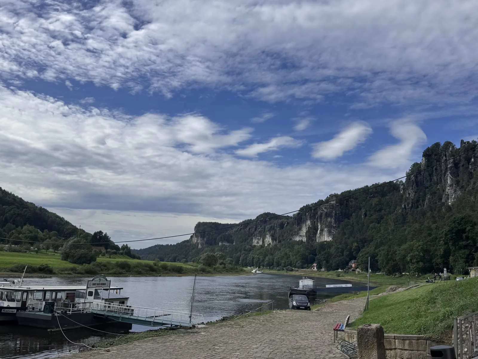 The Elbe running calm between forested sandstone cliffs, with a small ferry moored at a landing under a wide blue sky with scattered clouds.