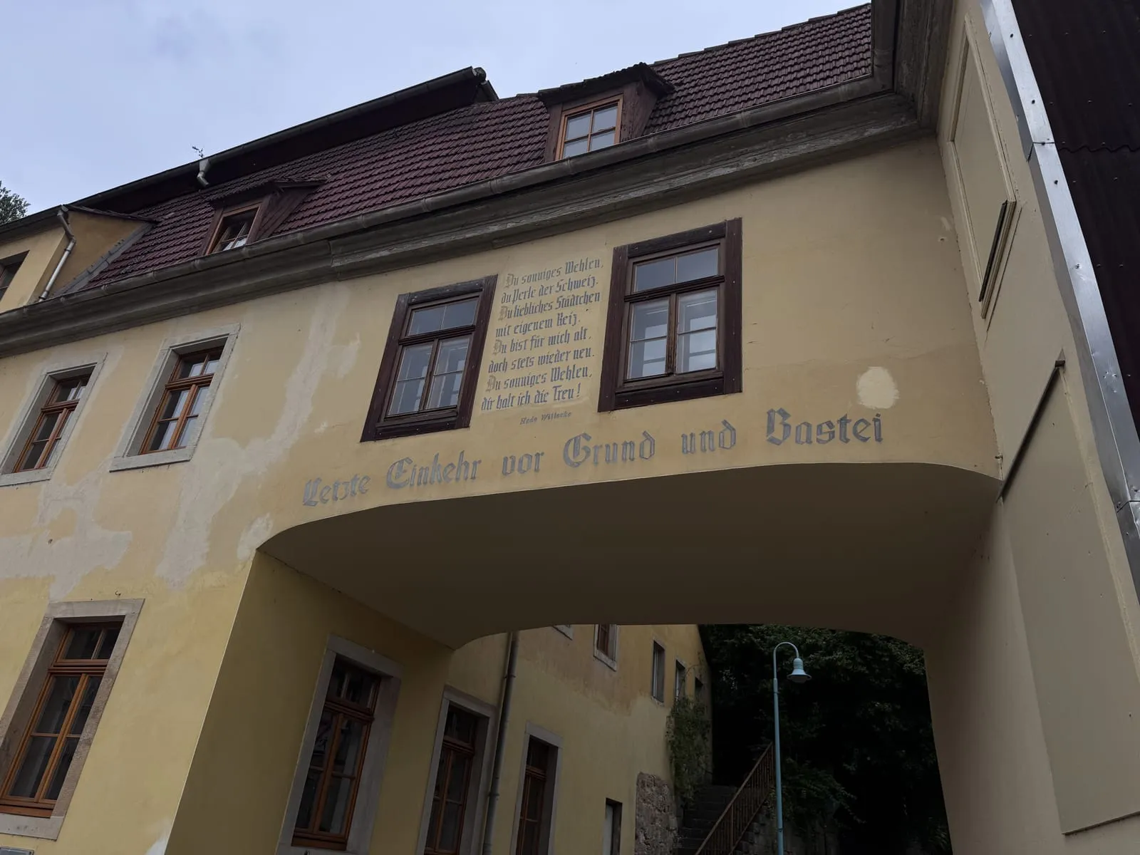 A yellow house arching over a passage, painted with the words Letzte Einkehr vor Grund und Bastei and a verse in praise of Wehlen.